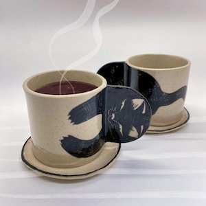 May include: Two ceramic mugs with matching saucers. The mugs are off-white with a speckled texture and feature a black cat design on the handle. One mug contains a dark liquid, with steam rising. The saucers are the same off-white color.