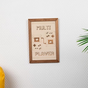 Peut inclure: Une impression encadrée avec un cadre en bois et le texte "MULTI PLAYER" avec un graphique de deux manettes de jeu vidéo.