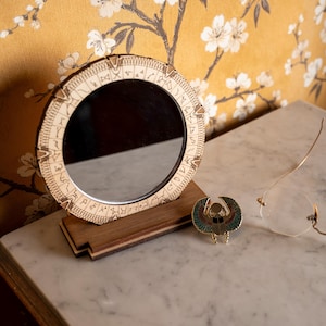 Könnte beinhalten: Ein runder Schminkspiegel aus Holz mit geätzten Symbolen und einer dunklen, reflektierenden Oberfläche. Eine kleine, goldfarbene Skarabäus-Anstecknadel mit blauen, roten und grünen Akzenten liegt daneben. Eine goldgerahmte Brille ist ebenfalls vorhanden.