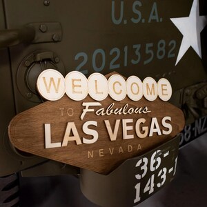 May include: Wooden sign that says "WELCOME to Fabulous LAS VEGAS NEVADA" attached to a military vehicle. The sign is brown with white lettering. The vehicle is olive green with white lettering and a white star.
