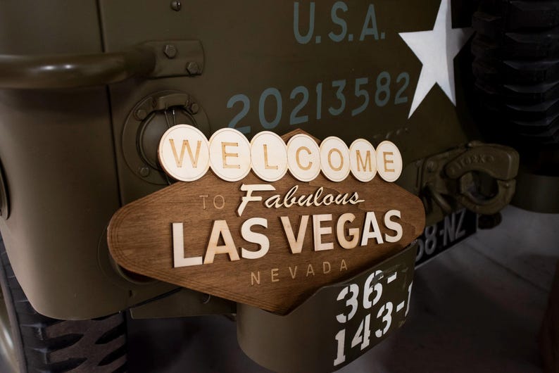 May include: Wooden sign that says "WELCOME to Fabulous LAS VEGAS NEVADA" attached to a military vehicle. The sign is brown with white lettering. The vehicle is olive green with white lettering and a white star.