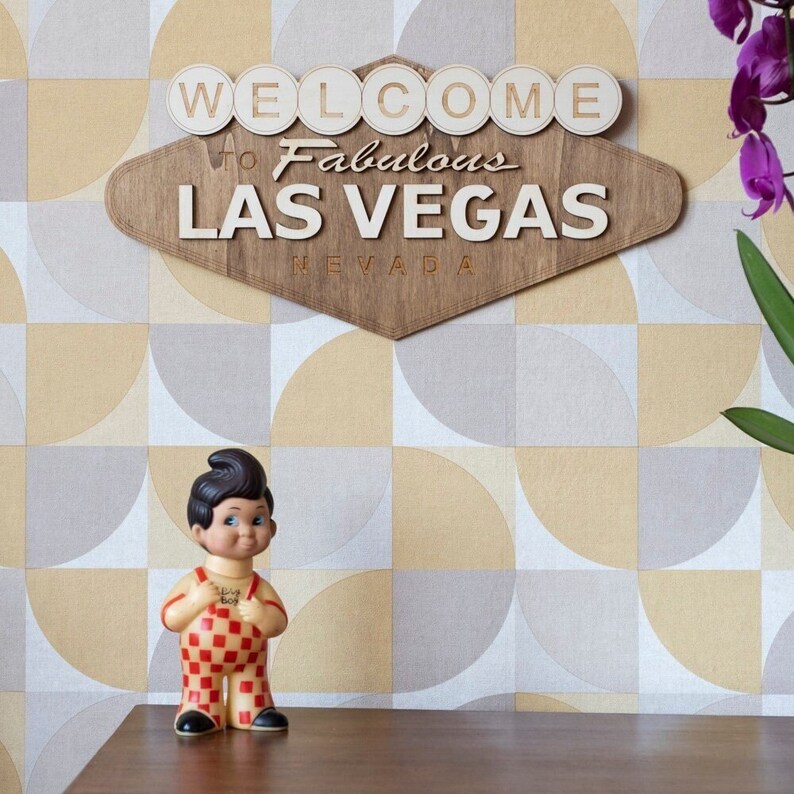 May include: Wooden sign with the words "WELCOME to Fabulous LAS VEGAS NEVADA". A vintage Big Boy figurine stands on a wooden surface in front of a geometric patterned wall. The sign is brown with white lettering.