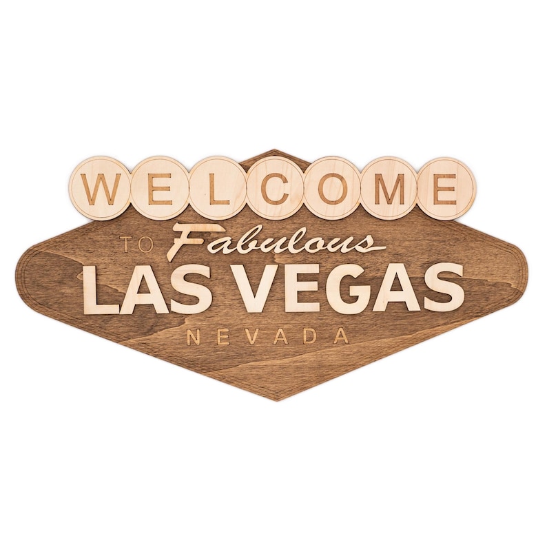 May include: Wooden sign with the words "WELCOME TO Fabulous LAS VEGAS NEVADA". The sign has a diamond shape with a dark brown stain and light brown lettering. The word "WELCOME" is in individual circles.