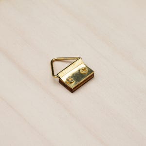 May include: A brass picture hanger with a rectangular wooden base and two screws. The hanger has a triangular loop for hanging and is designed to be mounted on a wooden surface. The hardware is gold-toned.