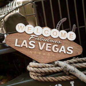 May include: Wooden sign with the words "WELCOME to Fabulous LAS VEGAS NEVADA" in white lettering. The sign is brown and diamond-shaped, with white circular letters above. A thick rope is in the foreground, and the sign is mounted on a military vehicle.