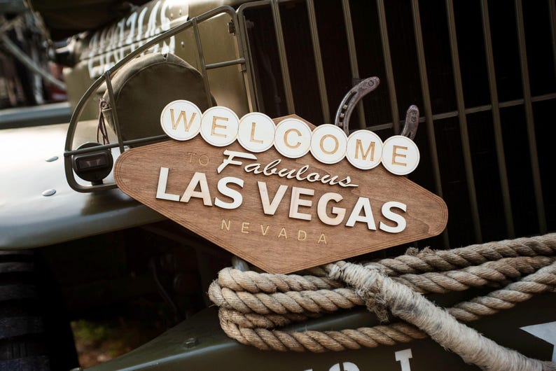 May include: Wooden sign with the words "WELCOME to Fabulous LAS VEGAS NEVADA" in white lettering. The sign is brown and diamond-shaped, with white circular letters above. A thick rope is in the foreground, and the sign is mounted on a military vehicle.
