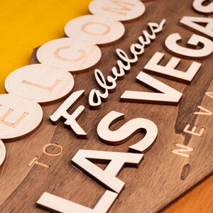 May include: Wooden sign with the words "Welcome to Fabulous Las Vegas Nevada" in a mix of fonts and styles. The sign is made of dark stained wood with light-colored wooden letters. The background is a bright yellow.
