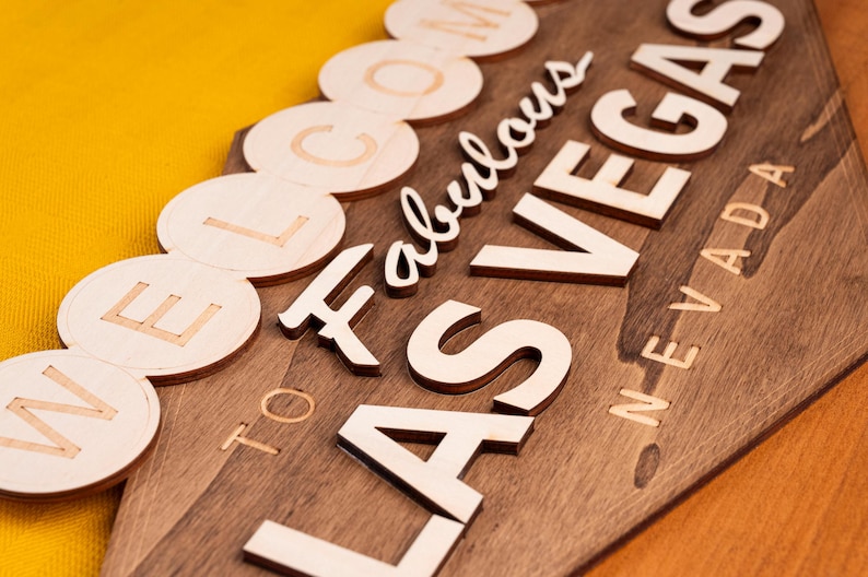 May include: Wooden sign with the words "Welcome to Fabulous Las Vegas Nevada" in a mix of fonts and styles. The sign is made of dark stained wood with light-colored wooden letters. The background is a bright yellow.
