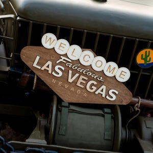 May include: A wooden sign with the text "WELCOME to Fabulous LAS VEGAS NEVADA" is mounted on the front of a military vehicle. The sign is brown with white lettering and circular accents. A cactus logo is visible.