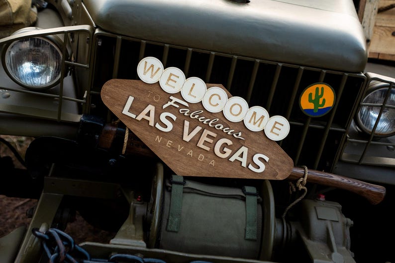 May include: A wooden sign with the text "WELCOME to Fabulous LAS VEGAS NEVADA" is mounted on the front of a military vehicle. The sign is brown with white lettering and circular accents. A cactus logo is visible.