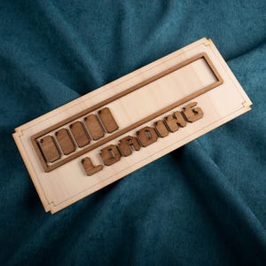 May include: Wooden sign with the word "LOADING" in pixelated font. The sign has a rectangular frame and a progress bar design. The sign is made of light-colored wood with darker brown accents.