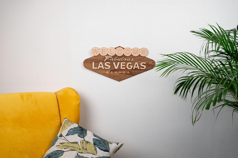May include: Wooden Las Vegas sign with the text "Welcome to Fabulous Las Vegas Nevada". The sign is brown with light-colored wooden letters and is mounted on a white wall. A yellow chair and a pillow with a floral pattern are in the foreground.