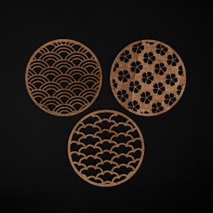 Puede incluir: Tres posavasos redondos de madera con intrincados diseños recortados. Uno presenta un patrón de olas, otro un diseño floral y el tercero un patrón de nubes. Los posavasos son de color marrón cálido y están sobre un fondo negro.