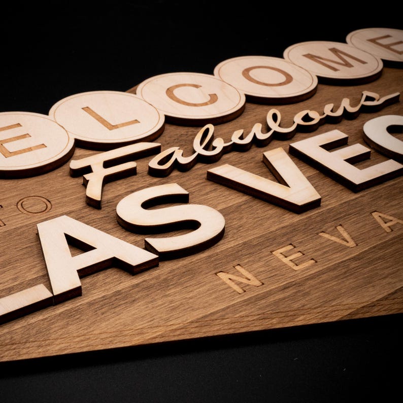 May include: Wooden sign with the words "Welcome Fabulous Las Vegas Nevada" in a mix of fonts and styles. The sign is made of light-colored wood with a dark wood grain pattern.