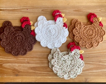 Pode incluir: Quatro tapetes de crochê em forma de frango em marrom, branco, bege e verde. Os tapetes são feitos com um fio texturizado e têm uma crista vermelha e amarela.