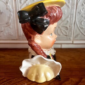 Pu&ograve; includere: Una statuetta in ceramica di una ragazza con un cappello giallo con un fiocco nero e un vestito bianco. La statuetta &egrave; un vaso di fiori in stile vintage.