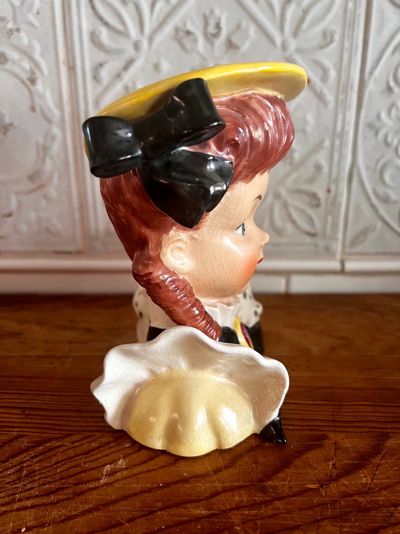 Pu&ograve; includere: Una statuetta in ceramica di una ragazza con un cappello giallo con un fiocco nero e un vestito bianco. La statuetta &egrave; un vaso di fiori in stile vintage.