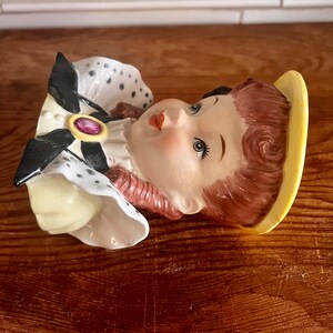 Pu&ograve; includere: Una statuetta in ceramica di una testa di donna con un'aureola gialla, un vestito a pois neri e bianchi e un gioiello rosa. La statuetta &egrave; un vaso da fiori in stile vintage.