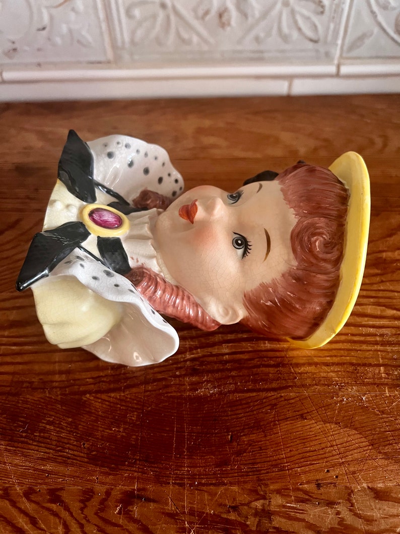 Pu&ograve; includere: Una statuetta in ceramica di una testa di donna con un'aureola gialla, un vestito a pois neri e bianchi e un gioiello rosa. La statuetta &egrave; un vaso da fiori in stile vintage.
