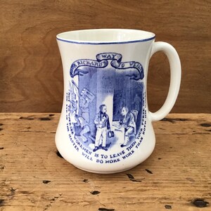 May include: A white ceramic mug with a blue and white design. The mug features a quote from Poor Richard's Almanack: "The Way to Wealth." The quote is surrounded by an illustration of a master and his apprentice.