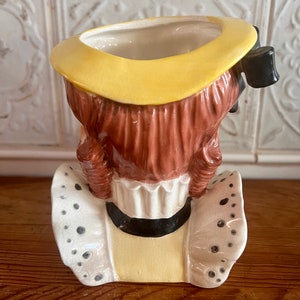 Pu&ograve; includere: Un vaso da fiori in ceramica a forma di ragazza con un cappello giallo e un colletto di pizzo bianco. Il vaso ha i capelli castani e un fiocco nero.