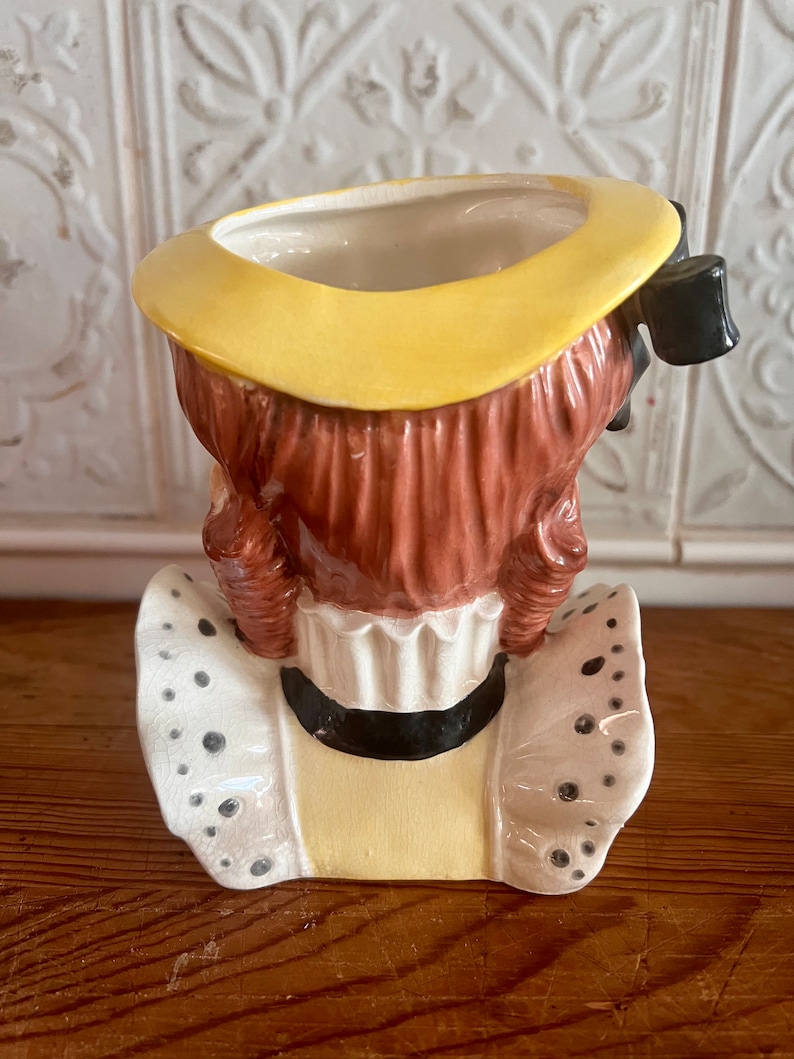 Pu&ograve; includere: Un vaso da fiori in ceramica a forma di ragazza con un cappello giallo e un colletto di pizzo bianco. Il vaso ha i capelli castani e un fiocco nero.