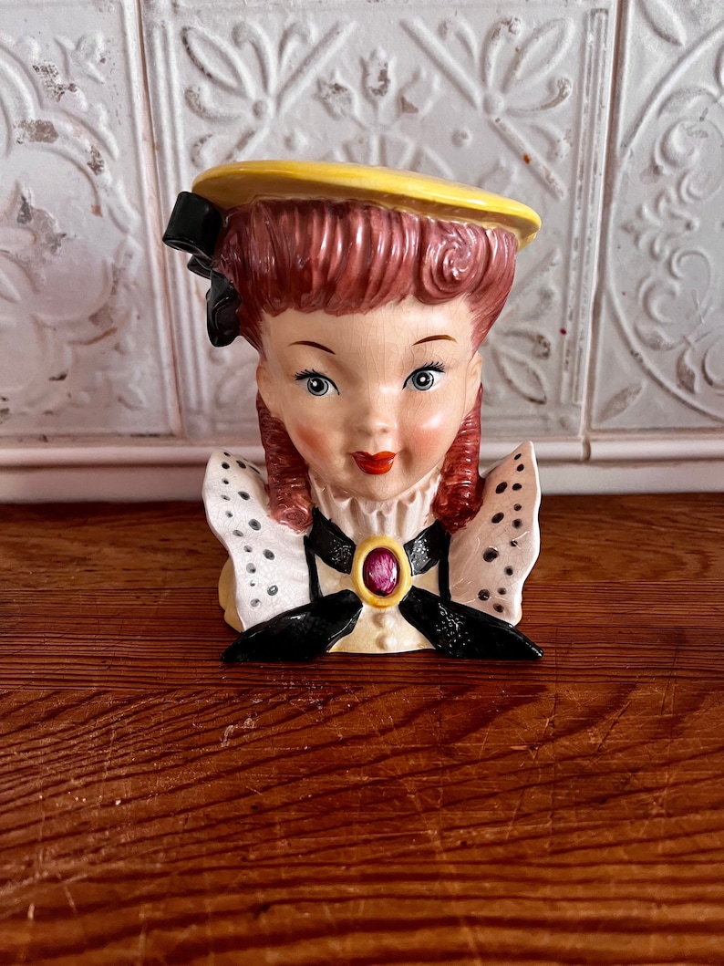 Pu&ograve; includere: Un busto di donna vintage in ceramica con un cappello giallo, un fiocco nero e un colletto a pois bianchi e neri. Il busto ha un viso rosa e bianco con una bocca rosa e occhi verdi. Il busto &egrave; un vaso con un foro sul retro per i fiori.