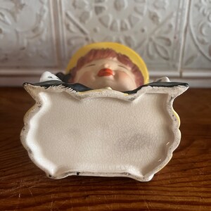 Pu&ograve; includere: Un vaso da fiori in ceramica bianca a forma di ragazza con un cappello giallo e un vestito bianco e nero. Il vaso ha una smaltatura craquel&eacute; ed &egrave; mostrato dal basso.