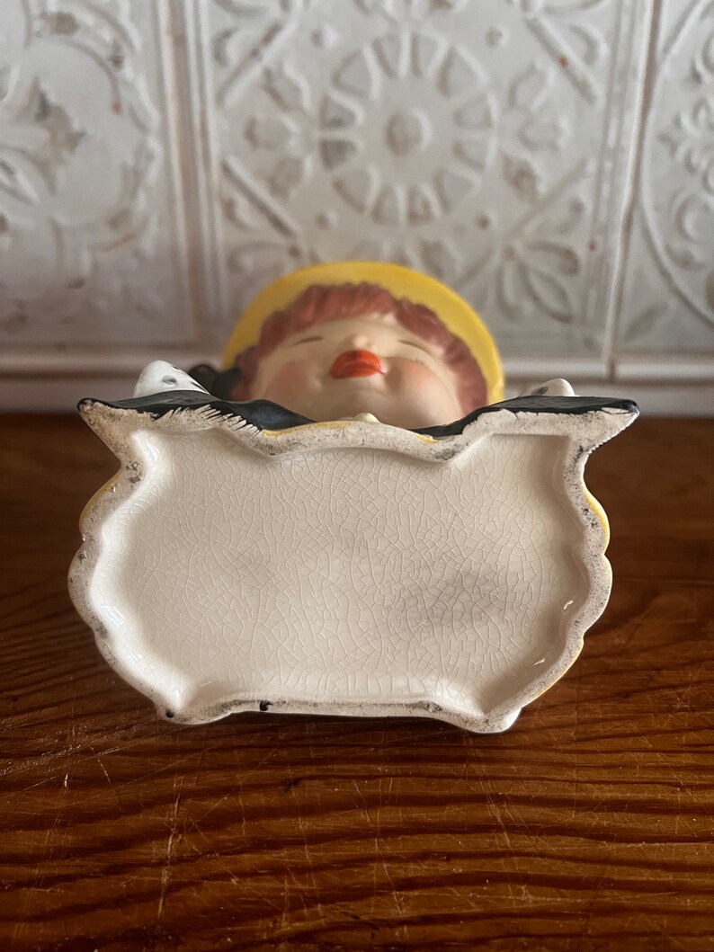 Pu&ograve; includere: Un vaso da fiori in ceramica bianca a forma di ragazza con un cappello giallo e un vestito bianco e nero. Il vaso ha una smaltatura craquel&eacute; ed &egrave; mostrato dal basso.
