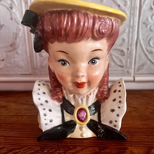 Pu&ograve; includere: Una statuetta in ceramica vintage di una testa di donna con un cappello giallo, un fiocco nero e un colletto a pois bianchi e neri. La statuetta ha un viso rosa e bianco con una bocca rossa e un gioiello viola sul colletto.