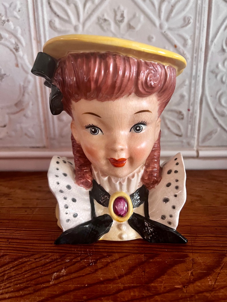Pu&ograve; includere: Una statuetta in ceramica vintage di una testa di donna con un cappello giallo, un fiocco nero e un colletto a pois bianchi e neri. La statuetta ha un viso rosa e bianco con una bocca rossa e un gioiello viola sul colletto.