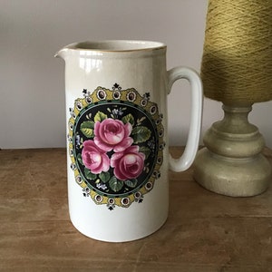 May include: White ceramic pitcher with a floral design. The design features pink roses, green leaves, and black and yellow accents. The pitcher has a gold rim and a handle.