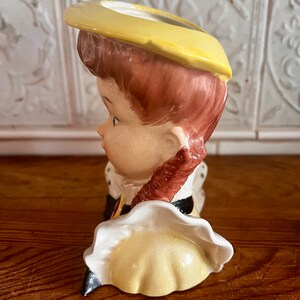 Pu&ograve; includere: Una statuetta in ceramica di una ragazza con i capelli castani che indossa un cappello e un vestito gialli. La statuetta &egrave; un vaso con un foro nella parte superiore per i fiori.