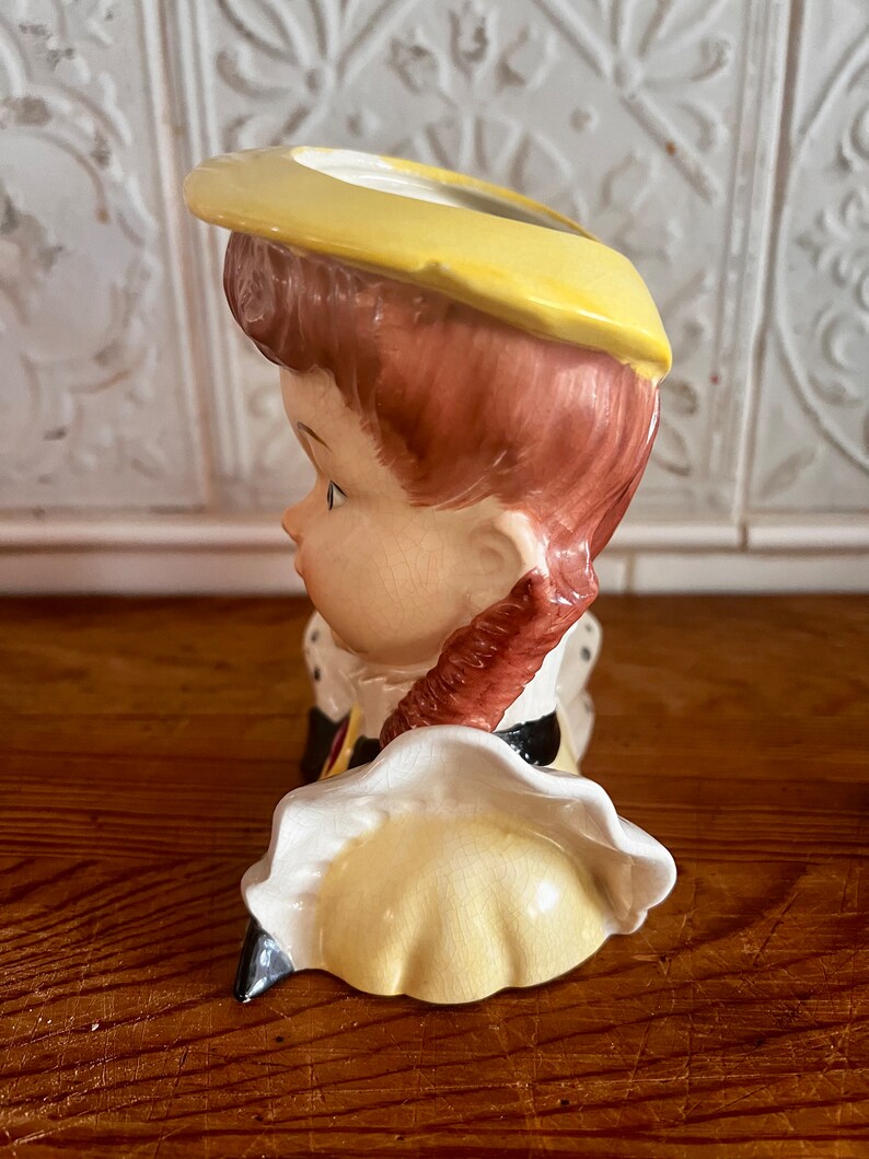 Pu&ograve; includere: Una statuetta in ceramica di una ragazza con i capelli castani che indossa un cappello e un vestito gialli. La statuetta &egrave; un vaso con un foro nella parte superiore per i fiori.