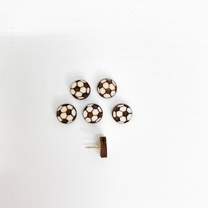 Puede incluir: Seis chinchetas de madera con forma de bal&oacute;n de f&uacute;tbol sobre un fondo blanco. Las chinchetas son marrones con un dise&ntilde;o de bal&oacute;n de f&uacute;tbol blanco. Hay una chincheta adicional con una cabeza dorada.