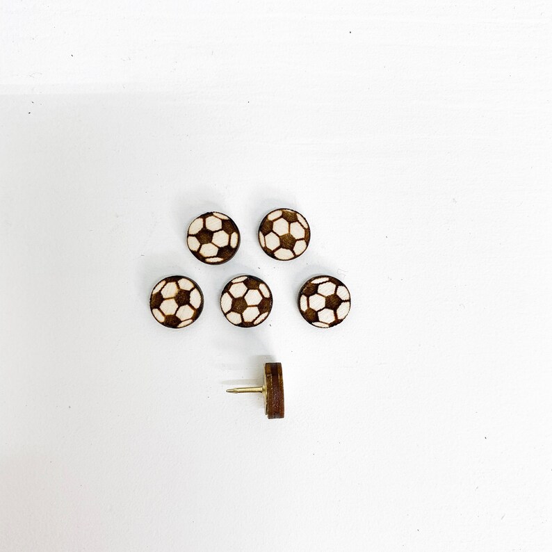Puede incluir: Seis chinchetas de madera con forma de bal&oacute;n de f&uacute;tbol sobre un fondo blanco. Las chinchetas son marrones con un dise&ntilde;o de bal&oacute;n de f&uacute;tbol blanco. Hay una chincheta adicional con una cabeza dorada.