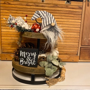 Puede incluir: Una bandeja de madera de dos niveles con un soporte de metal negro. La bandeja está decorada con un gnomo que lleva un gorro a rayas blanco y negro, un adorno rojo, piñas, vegetación y un letrero de madera que dice "Merry & Bright".
