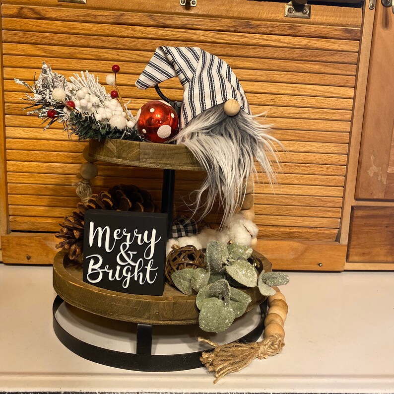 May include: A two-tier wooden tiered tray with a black metal stand. The tray is decorated with a gnome wearing a black and white striped hat, a red ornament, pine cones, greenery, and a wooden sign that says "Merry & Bright".