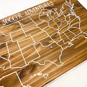 Puede incluir: Un mapa de madera de los Estados Unidos con l&iacute;neas de estado blancas y las palabras "Soccer Adventures" en la parte superior. Hay alfileres blancos en varios estados.