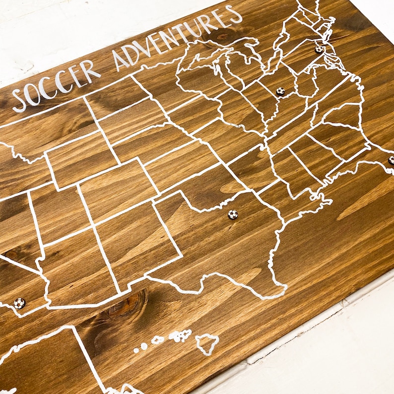 Puede incluir: Un mapa de madera de los Estados Unidos con l&iacute;neas de estado blancas y las palabras "Soccer Adventures" en la parte superior. Hay alfileres blancos en varios estados.