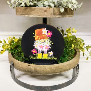 May include: A tiered wooden tray with a black painted round wooden gnome ornament. The gnome is wearing a plaid shirt, orange hat, and yellow boots. The gnome is surrounded by pink and white flowers.