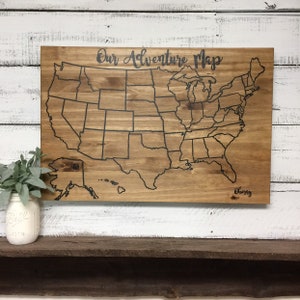 May include: Wooden adventure map of the United States with state outlines etched in black. The words "Our Adventure Map" are scripted across the top. A personalized name is written in cursive at the bottom right.
