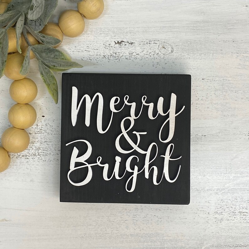 May include: Black wooden sign with white lettering that reads "Merry & Bright".