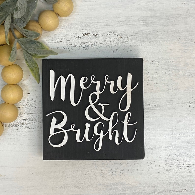 May include: Black wooden sign with white lettering that reads "Merry & Bright".