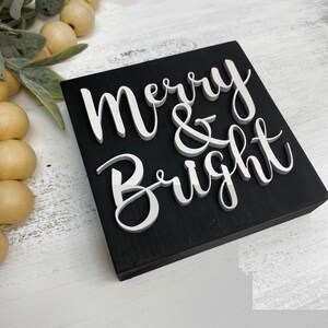 May include: Black wooden sign with white raised lettering that reads "Merry & Bright".