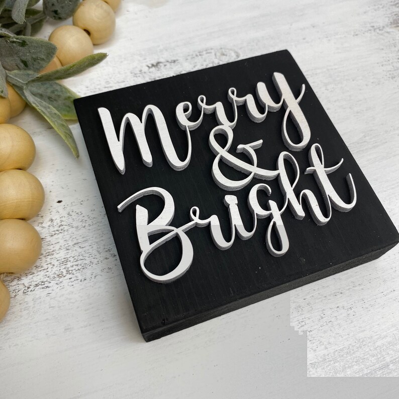 May include: Black wooden sign with white raised lettering that reads "Merry & Bright".
