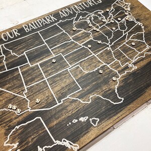 Puede incluir: Un mapa de madera de los Estados Unidos con el texto "Our Ballpark Adventures" en la parte superior. Se colocan alfileres con forma de pelota de b&eacute;isbol en el mapa para marcar ubicaciones.