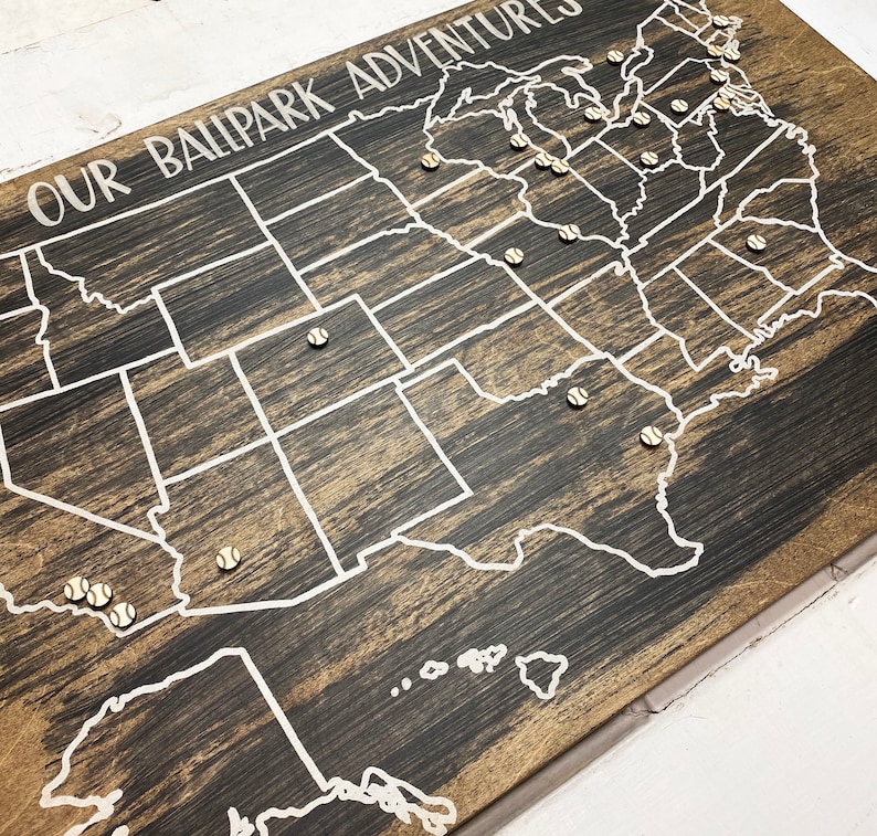 Puede incluir: Un mapa de madera de los Estados Unidos con el texto "Our Ballpark Adventures" en la parte superior. Se colocan alfileres con forma de pelota de b&eacute;isbol en el mapa para marcar ubicaciones.