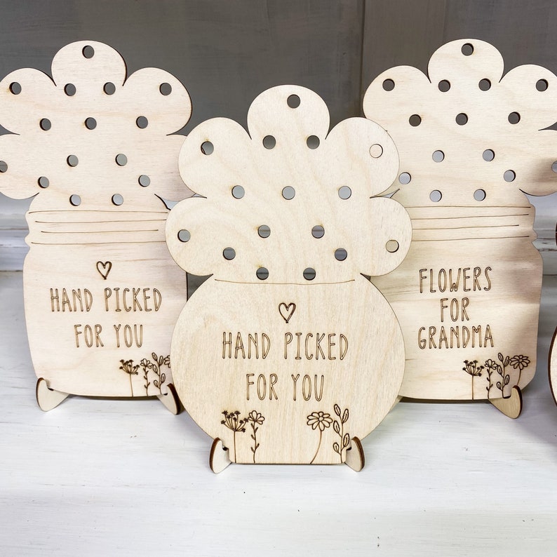 May include: Three wooden flower pot decorations with a cutout design of flowers. The pots have a message engraved on them: "Hand Picked For You", "Hand Picked For You", and "Flowers For Grandma".