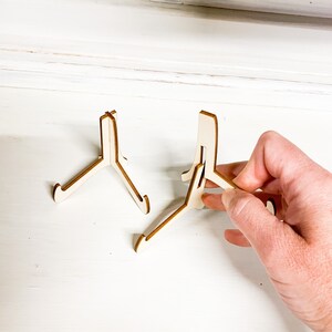 May include: Two wooden picture stands, one is folded and the other is being unfolded by a hand.